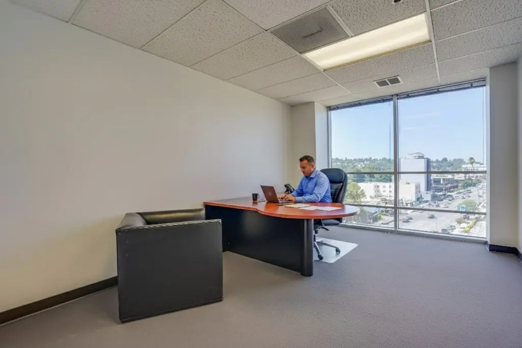Barrister Suites Office Lease A man in a blue shirt sits alone at a desk in a spacious, modern office with large windows and a city view, working on a laptop. An empty black chair is in front of the desk.