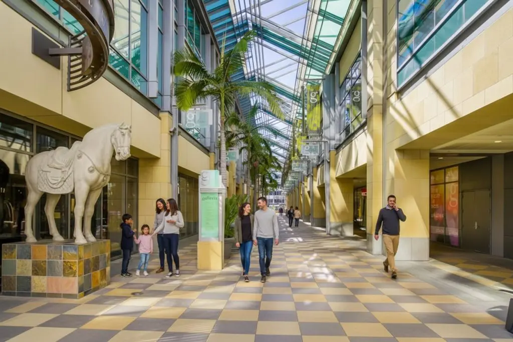 Barrister Suites Office Lease People walk and talk inside a bright, modern shopping mall with glass ceilings, palm trees, banners, and a white horse statue. Families and individuals enjoy the spacious, sunlit corridor.