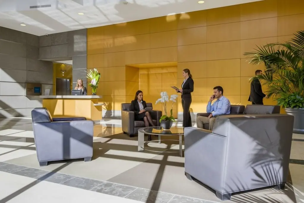 Barrister Suites Office Lease Modern office lobby with large windows, people sitting on black chairs, using phones or tablets, and a receptionist behind a wooden desk. Light streams in, plants and flowers decorate the space.