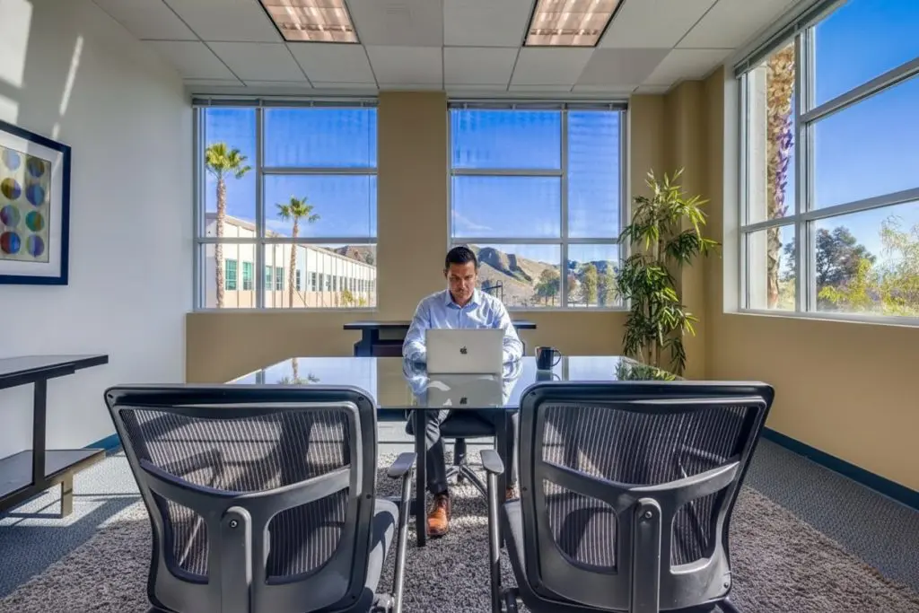 Barrister Suites Office Lease A man in business attire sits at a desk working on a laptop in a modern office with large windows, two empty chairs in front, and a scenic view of mountains and palm trees outside.