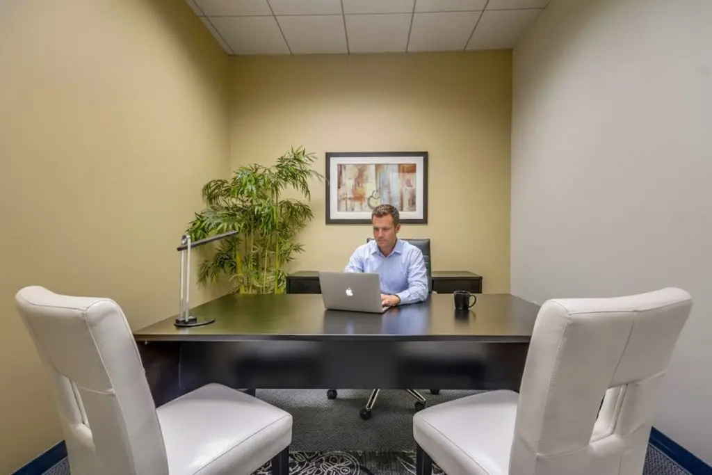 Barrister Suites Office Lease A man sits alone at a desk in a small office, working on a laptop. Two white chairs face his desk, a mug and desk lamp are on the table, and a potted plant and framed art hang on the beige walls.