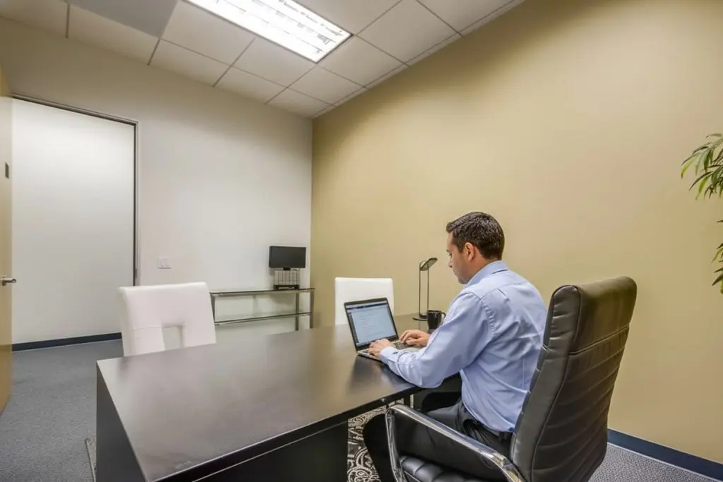 Barrister Suites Office Lease A man in a blue shirt sits at a desk in a modern office, working on a laptop. The room has beige walls, white chairs, a small table with a lamp, and a plant in the corner.