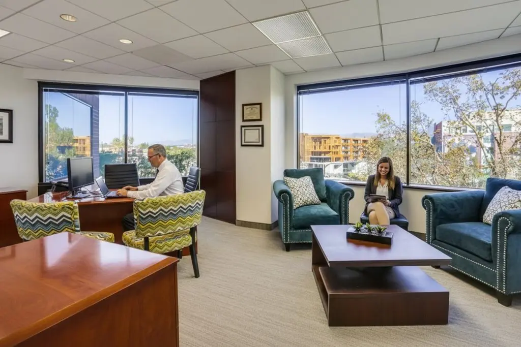 Barrister Suites Office Lease A modern office with large windows, a man working at a desk, and a woman sitting and reading on a blue armchair. There are two patterned chairs, a coffee table, and trees and buildings visible outside.