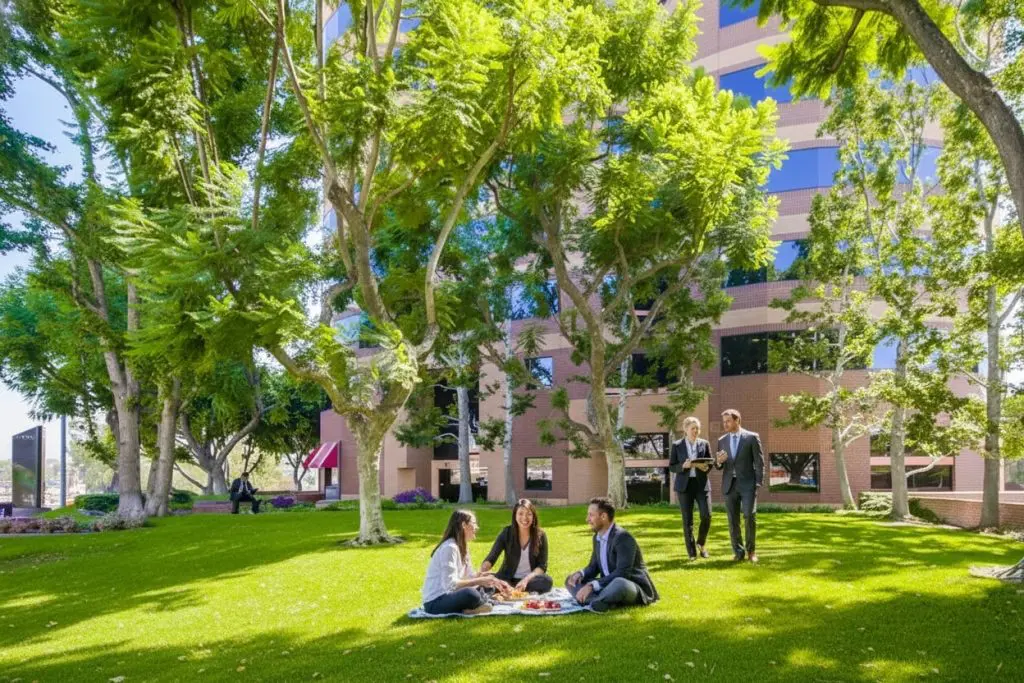 Barrister Suites Office Lease Four people sit on the grass having a picnic under trees in front of an office building, while three others in business attire walk nearby. Sunlight streams through the leaves, creating a bright and relaxed atmosphere.