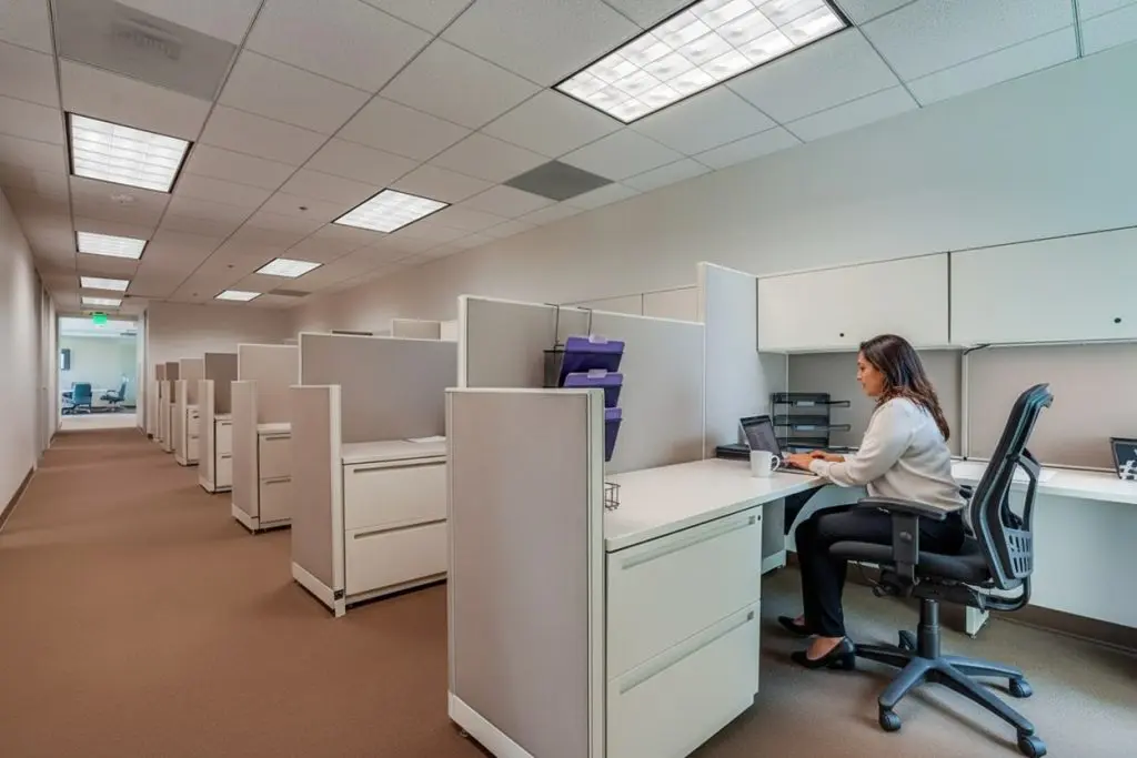 Barrister Suites Office Lease A woman sits alone at a desk in a modern, empty office cubicle area, working on a laptop. The office has beige carpeting, white cubicles, and overhead fluorescent lights.