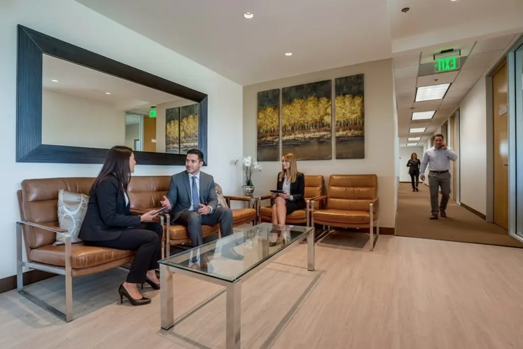 Barrister Suites Office Lease Four people are in a modern office lobby. Two people sit and talk on brown leather chairs, another sits nearby with a tablet, and a person walks down the hallway. Large art and a mirror decorate the room.