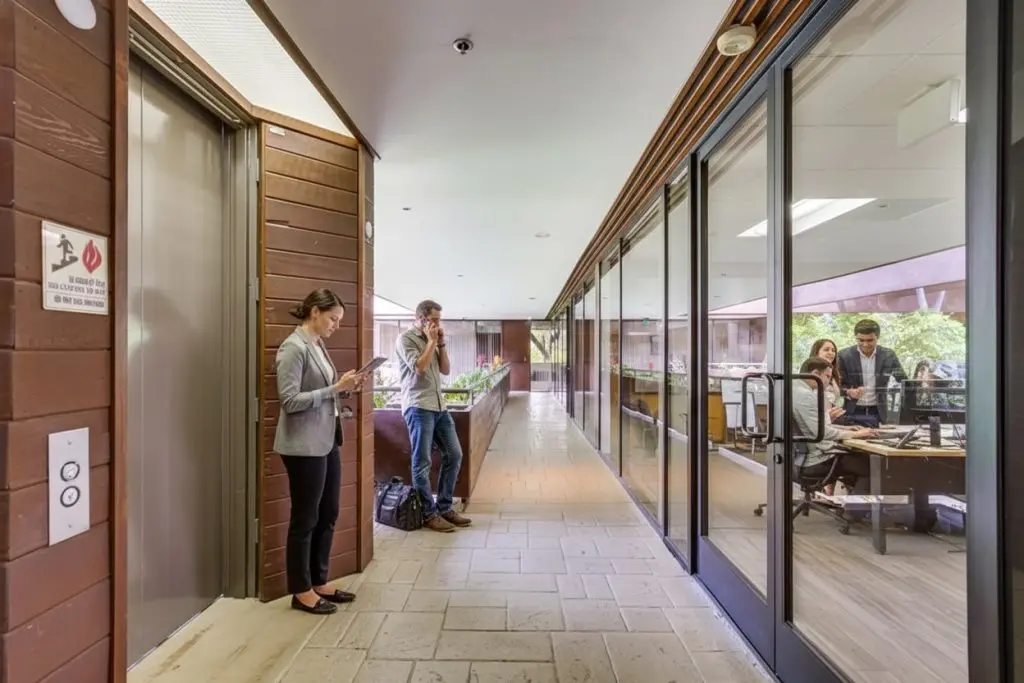 Barrister Suites Office Lease Two people stand in a hallway near an elevator, using their phones, while three colleagues work and talk at a conference table inside a glass-walled office to the right.