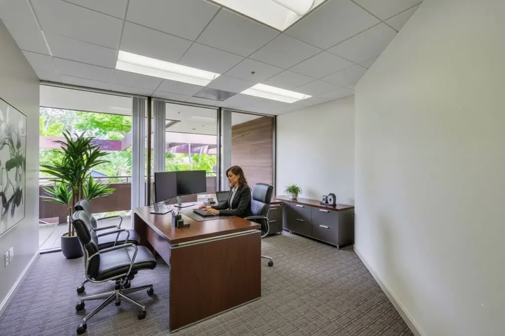 Barrister Suites Office Lease A woman in business attire sits at a wooden desk in a bright, modern office with large windows, two guest chairs, plants, and a computer monitor.