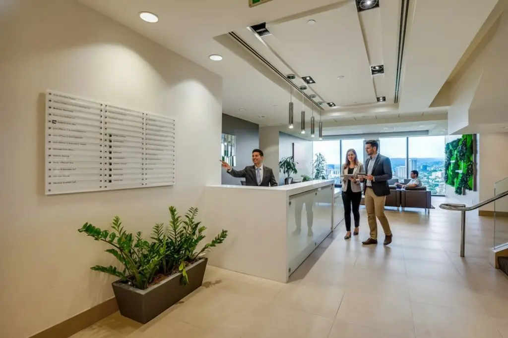 Barrister Suites Office Lease A receptionist gestures toward a wall directory as a man and woman approach the counter in a modern office lobby with plants, seating area, and large windows in the background.