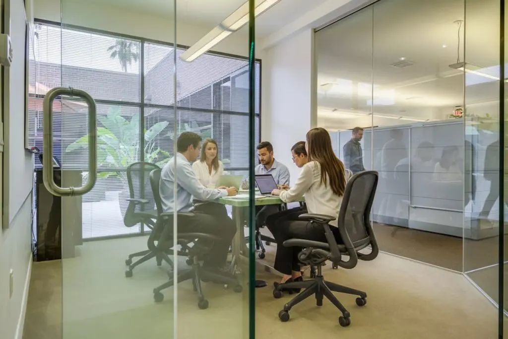 Barrister Suites Office Lease Five people in business attire sit around a table in a glass-walled office, engaged in a meeting. Laptops and notebooks are on the table. Bright, modern workspace with large windows and plants outside.