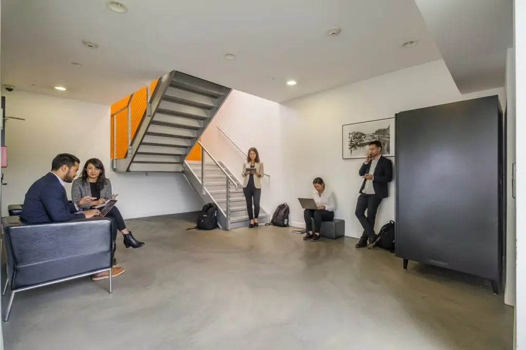 Barrister Suites Office Lease Five people in business attire are in a modern office lobby with gray couches and a staircase. Two sit on a couch using laptops, one sits on the floor with a laptop, and two stand using smartphones.