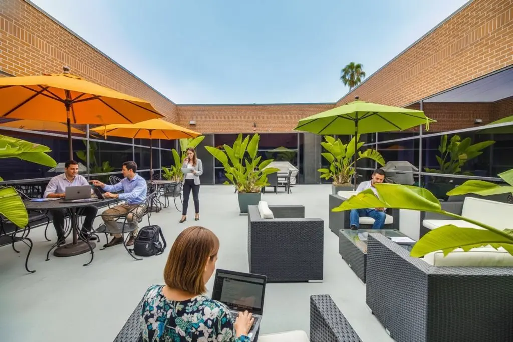 Barrister Suites Office Lease People work on laptops and talk on phones in an outdoor courtyard with patio furniture, large umbrellas, potted plants, and brick walls under a clear sky.