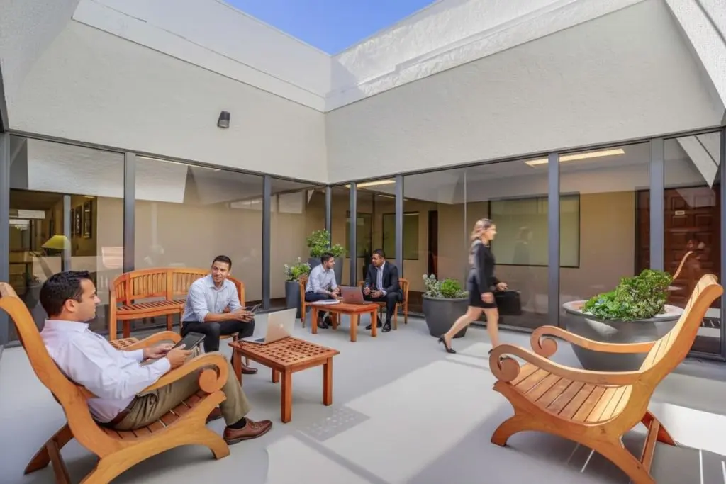 Barrister Suites Office Lease Four people sit and talk on wooden chairs in a modern outdoor courtyard, while one person walks by. Large windows, potted plants, and natural light create an open, professional atmosphere.