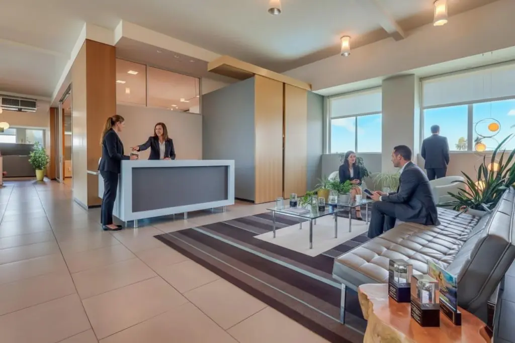 Barrister Suites Office Lease A modern office lobby with people in business attire; two women talk at a reception desk while three others sit and stand near large windows, surrounded by plants and contemporary furniture.