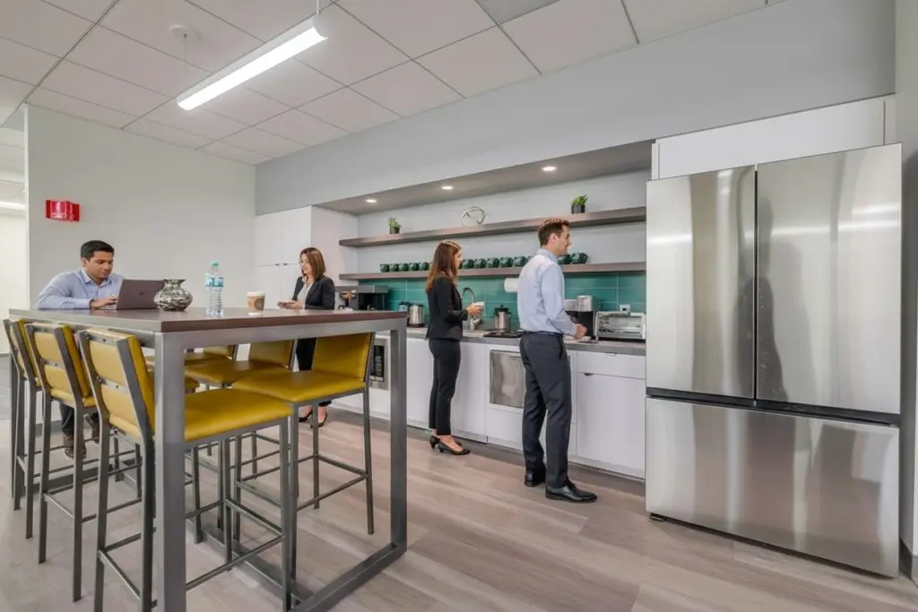 Barrister Suites Office Lease Four people are in a modern office kitchen; two are preparing drinks, one is using a laptop at a high table with yellow chairs, and another stands by the counter. The space is bright with stainless steel appliances.