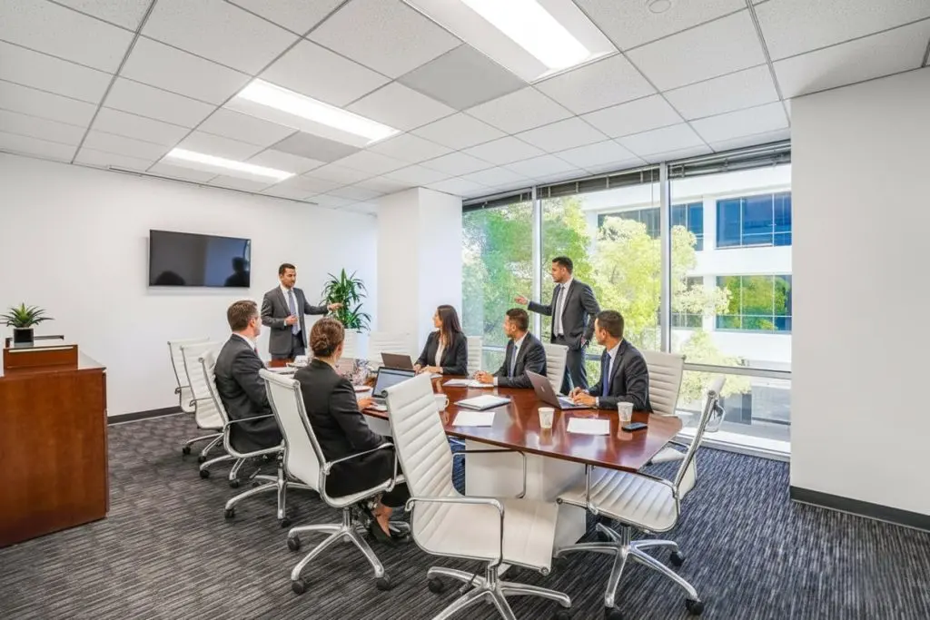 Barrister Suites Office Lease A group of seven people in business attire are having a meeting in a modern office with large windows. One man is standing and speaking, while others are seated around a table with laptops and documents.