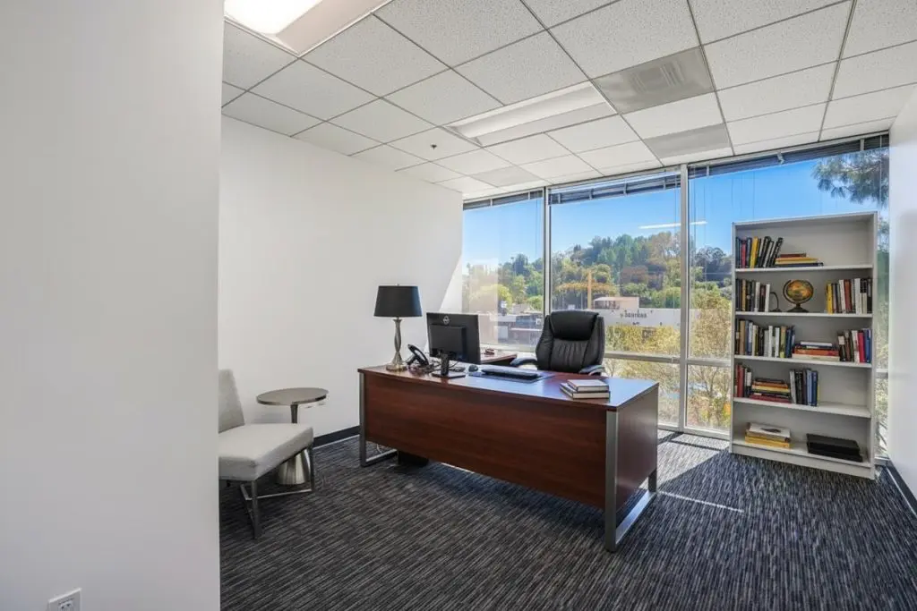 Barrister Suites Office Lease A modern office features a large desk with a black chair, lamp, and computer. There’s a gray chair and small table on the side, a bookshelf with books and decor, and large windows letting in natural light.