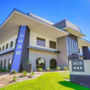 Barrister Suites Office Lease Modern two-story office building with a large sign displaying the number 3838. The building has a slanted roof, large windows, and landscaped greenery in front under a clear blue sky.