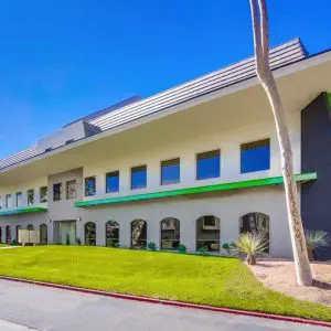 Barrister Suites Office Lease Modern two-story office building with large windows, green accents, and address number 3868 displayed vertically. The building is surrounded by a well-kept lawn and a few trees under a clear blue sky.