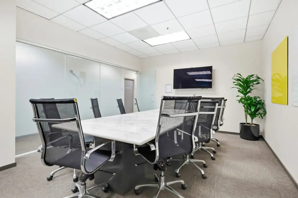 Barrister Suites Office Lease A modern conference room with a long marble table, eight black office chairs, a wall-mounted TV, a potted plant, white walls, abstract art, glass doors, and ceiling lights.