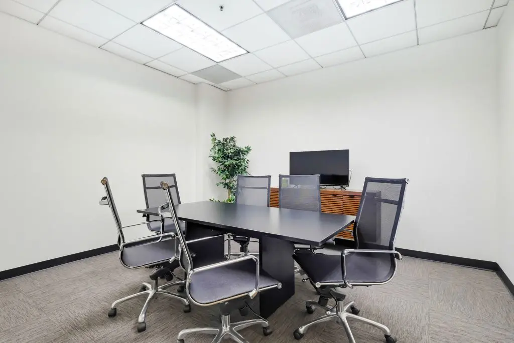 Barrister Suites Office Lease A small conference room with a black rectangular table, five mesh office chairs, a flat-screen monitor on a wooden cabinet, a potted plant in the corner, and white walls with a tiled ceiling.