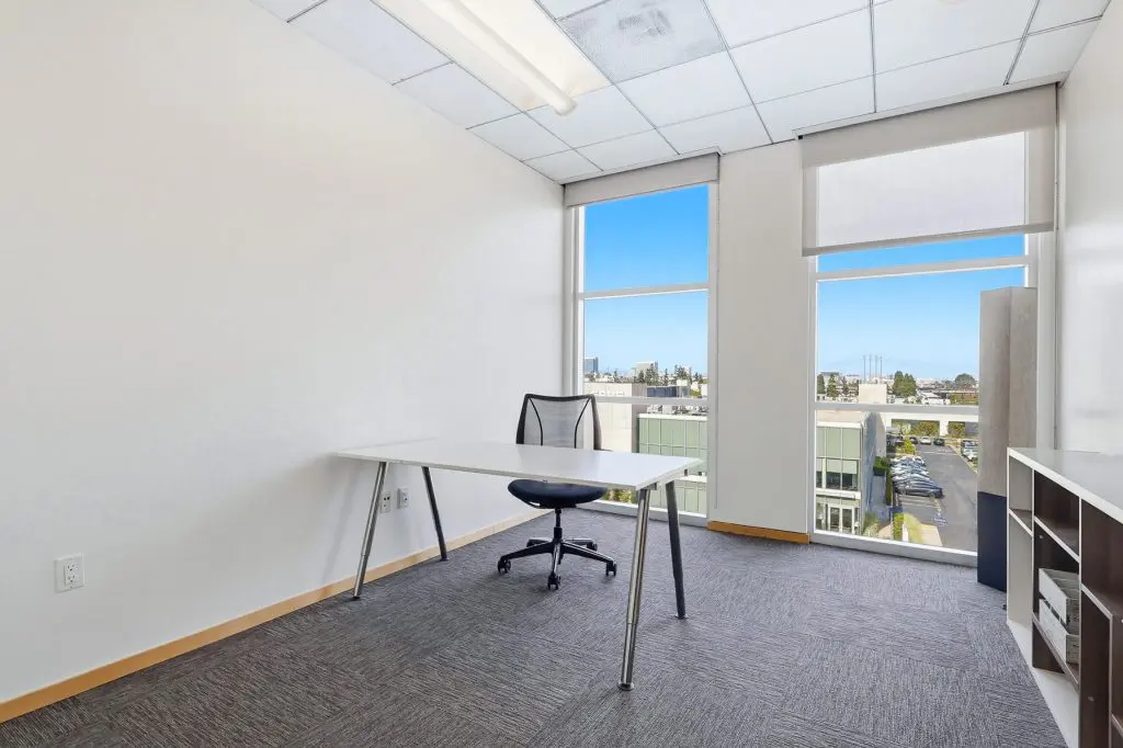 Barrister Suites Office Lease A modern, minimal office with a single desk, mesh chair, shelving unit, and large windows letting in natural light, overlooking city buildings and a blue sky.