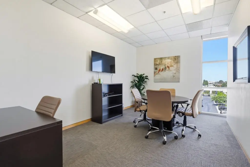 Barrister Suites Office Lease A modern office with beige chairs around a round table, a wall-mounted TV, a dark cabinet, a desk, a potted plant, and a large window letting in natural light.