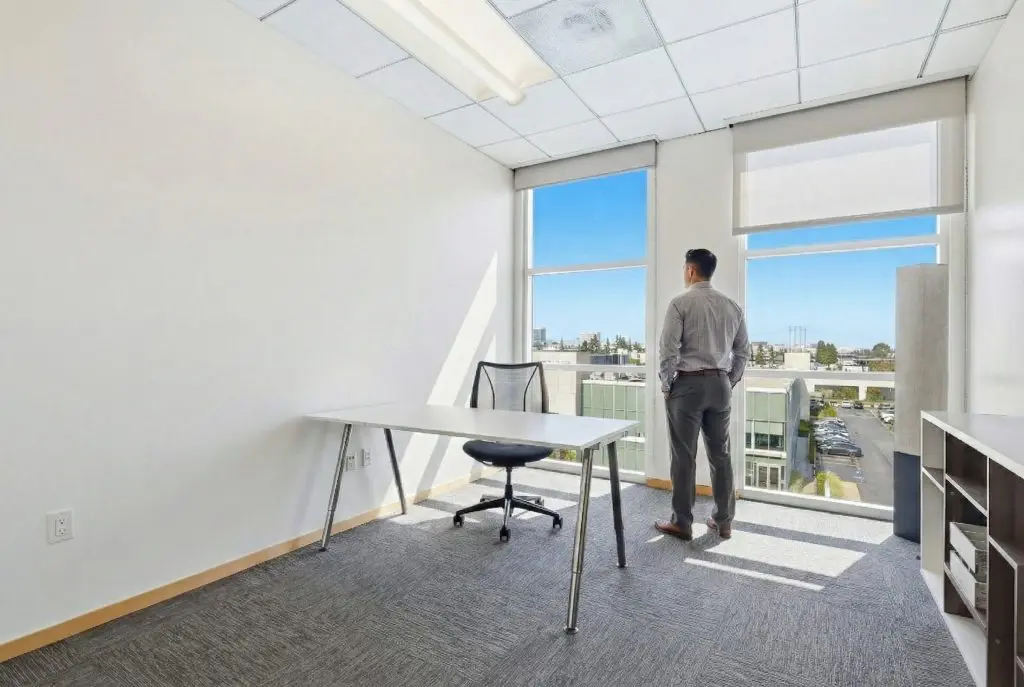Barrister Suites Office Lease A man in business attire stands by large windows in a bright, modern office, looking outside. The office has a desk, chair, shelving unit, and neutral decor. Sunlight fills the space, highlighting its clean, minimalist design.