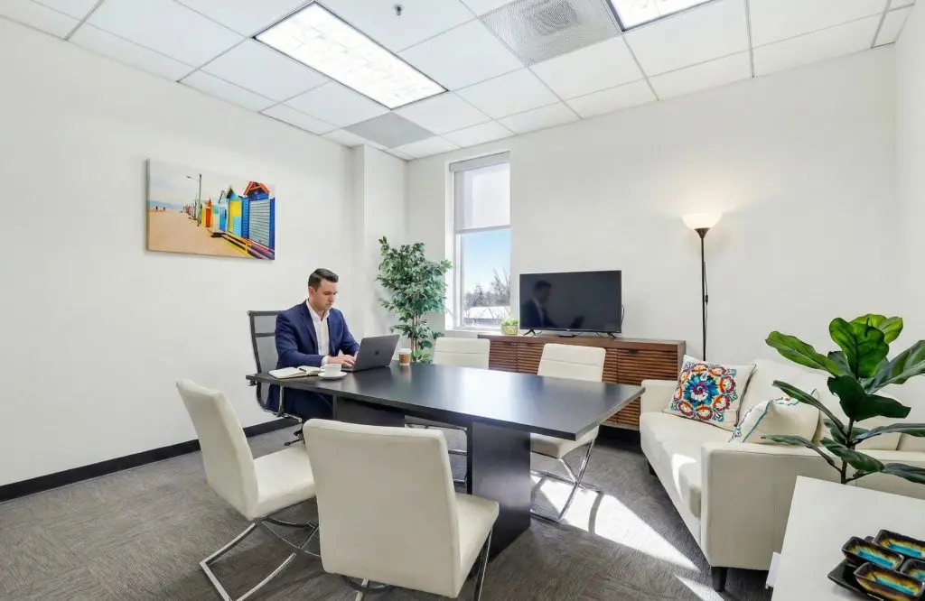 Barrister Suites Office Lease A man in a suit works on a laptop at a conference table in a modern, brightly lit office with white walls, a couch, plants, a TV, and colorful wall art.