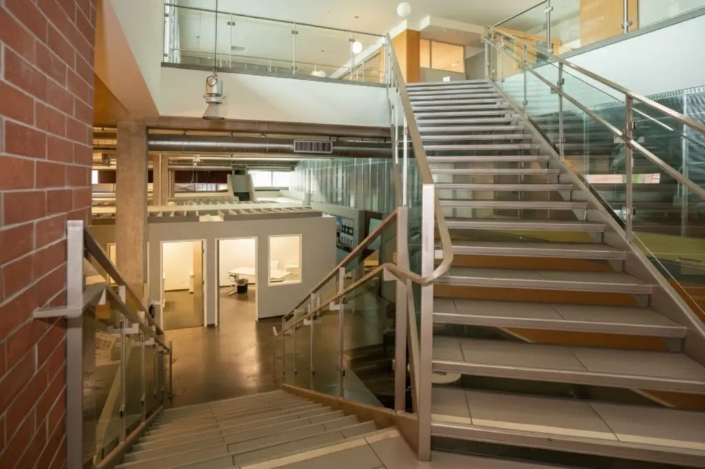 Barrister Suites Office Lease Modern indoor staircase with glass railings leads to an upper floor. The lower level shows open office spaces with lights on, white walls, and a brick wall on the left side. The area appears clean and well-lit.