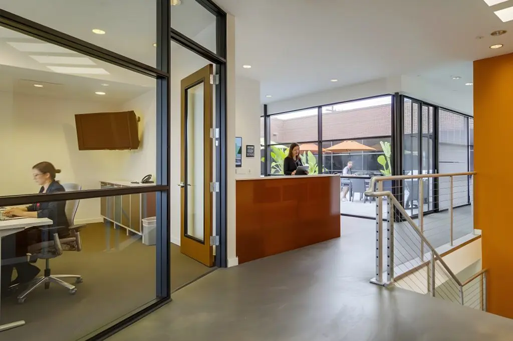 Barrister Suites Office Lease A modern office space with a glass-walled room on the left, a person working at a desk inside, and a reception area with another person at the counter. Large windows reveal an outdoor patio with tables and umbrellas.