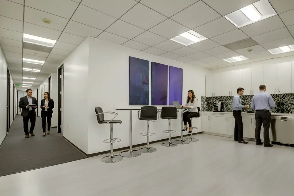 Barrister Suites Office Lease A modern office kitchen with white cabinets and bar stools. One woman sits at the counter using a laptop, while three men stand near the sink and coffee maker. Two people walk down a hallway to the left.