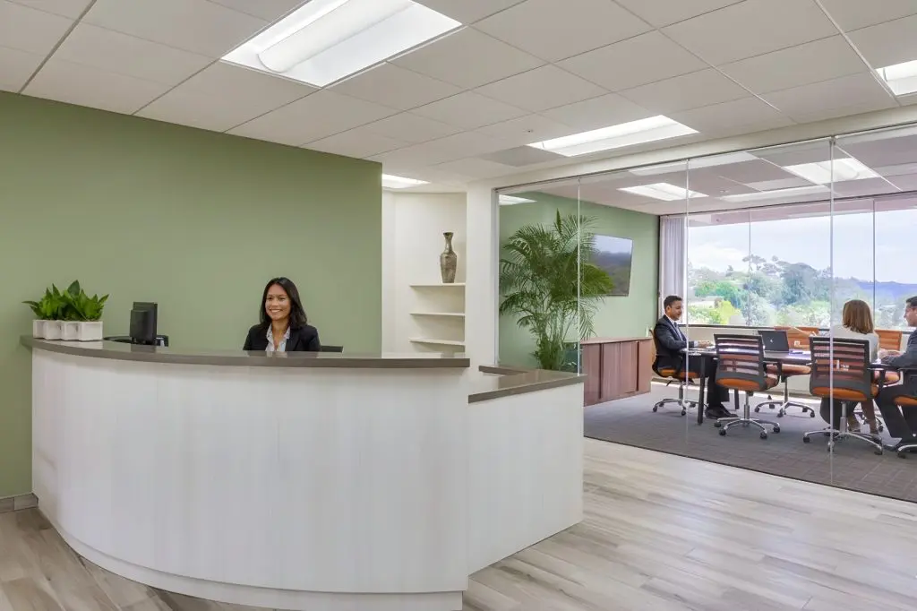 Barrister Suites Office Lease A modern office lobby with a smiling receptionist at a curved desk, green accent wall, and potted plants. Behind glass walls, several people are seated at a conference table in a meeting, with large windows showing a scenic outdoor view.