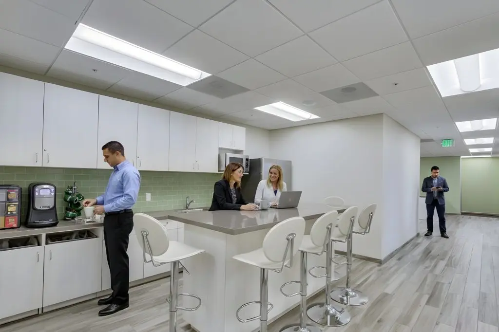 Barrister Suites Office Lease Modern office kitchen with white cabinets and a green backsplash. Three people talk at an island counter, one using a laptop, while another person makes coffee. A fourth person stands in the hallway looking at a tablet.