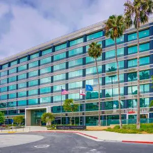 Barrister Suites Office Lease A modern glass office building with seven stories, palm trees in front, and three flags flying near the entrance under a cloudy sky. The street and some greenery are visible in the foreground.