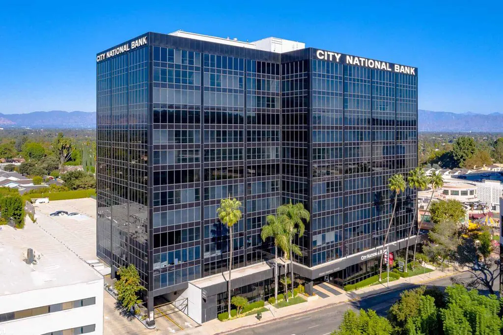 Barrister Suites Office Lease A tall, modern glass office building with City National Bank written at the top, surrounded by palm trees and other buildings under a clear blue sky.