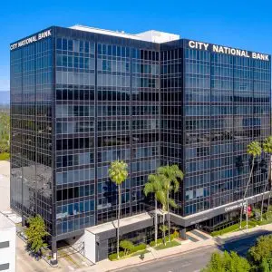 Barrister Suites Office Lease A tall, modern glass office building with City National Bank written at the top, surrounded by palm trees and other buildings under a clear blue sky.