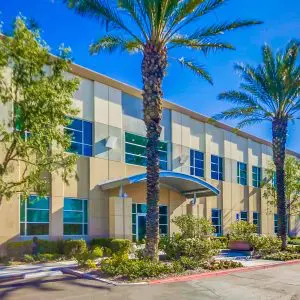 Barrister Suites Office Lease A modern two-story office building with large windows, palm trees, and shrubs in front, under a bright blue sky. A covered entrance is visible at the center.