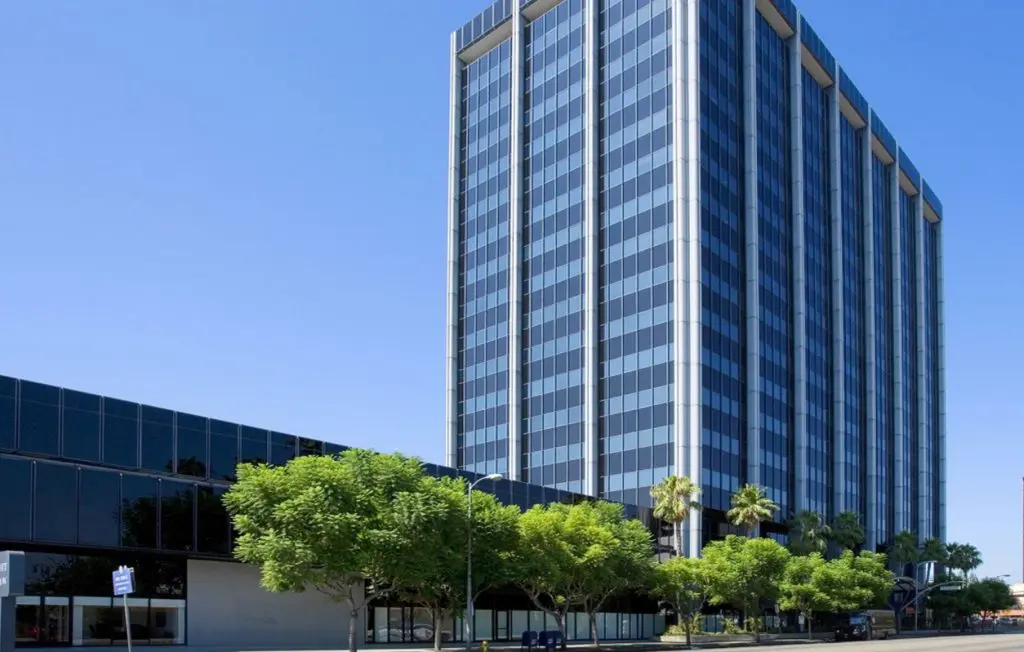 Barrister Suites Office Lease A tall, modern office building with mirrored glass windows stands next to a shorter building. Green trees line the sidewalk in front of the structures under a clear blue sky.