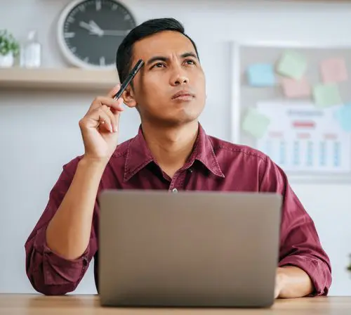 Barrister Suites Office Lease A man in a maroon shirt sits at a desk with a laptop, holding a pen to his temple and looking thoughtful. A clock and a corkboard with sticky notes are visible in the background.
