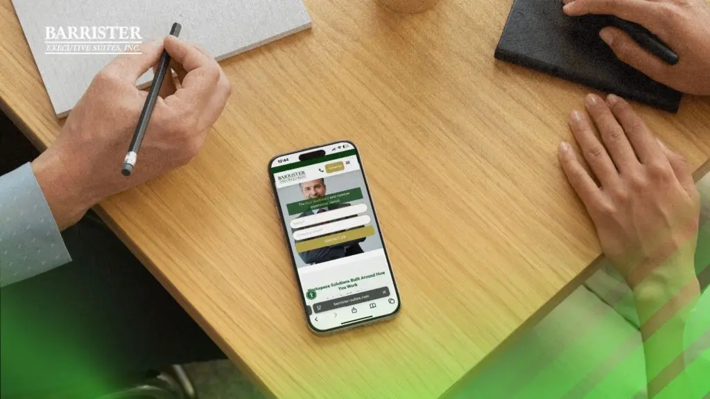 Barrister Suites Office Lease Two people sit at a wooden table with a phone displaying the Barrister Executive Suites website between them. One person holds a pencil; a notebook is nearby. The Barrister Executive Suites, Inc. logo is in the top left corner.