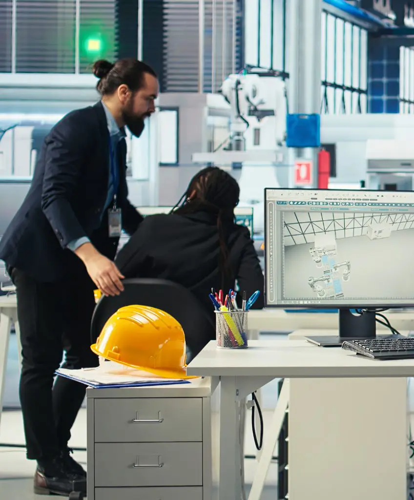 Barrister Suites Office Lease Two people in business attire work in a modern office with engineering software on a computer screen. A yellow hard hat and office supplies are on a nearby desk. The setting appears to be an industrial workspace.
