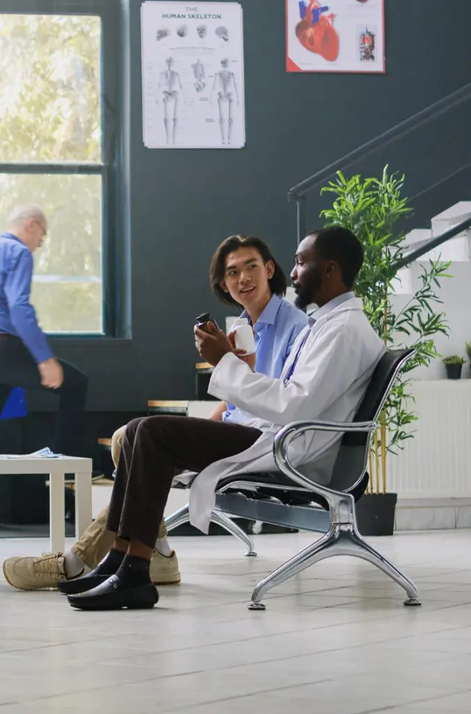 Barrister Suites Office Lease Two doctors sit and talk in a waiting area, one holding a phone and coffee cup. Medical posters and plants are visible in the background, and another person is seated near the window.