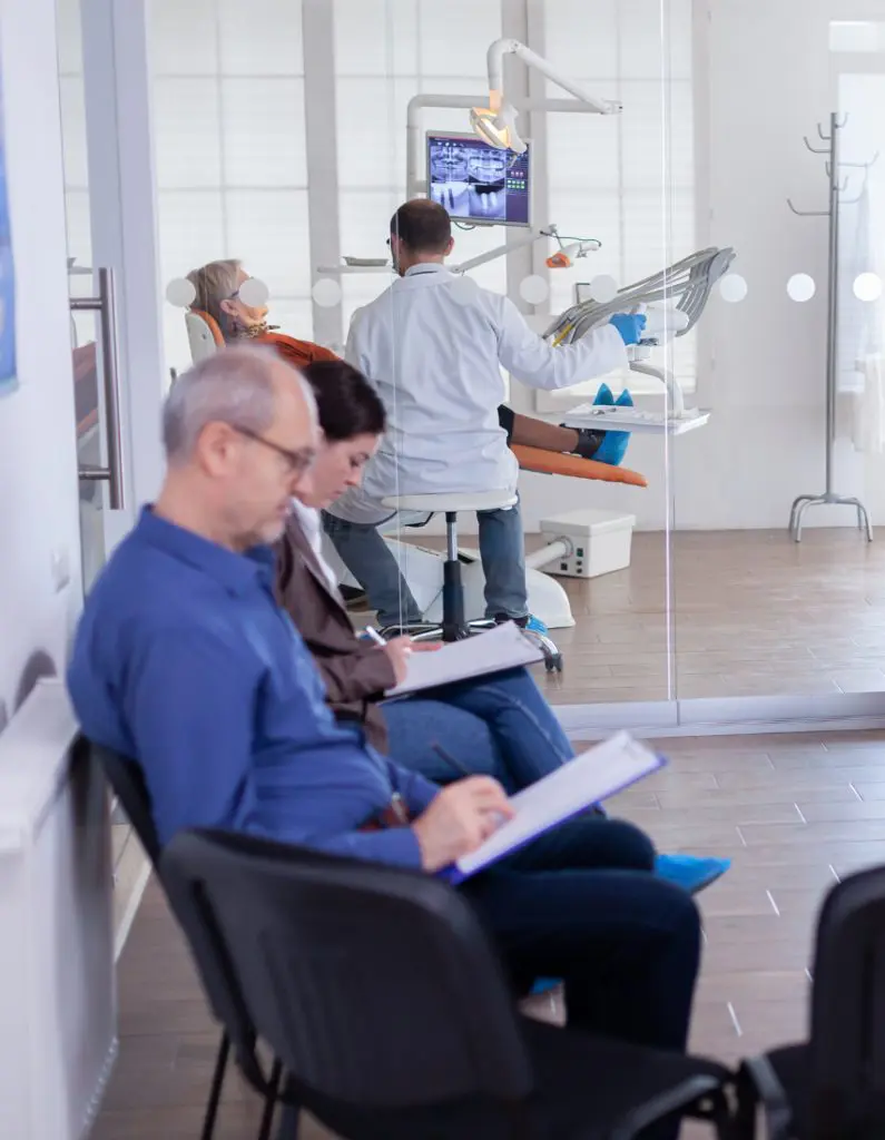 Barrister Suites Office Lease Two people sit in a waiting room filling out forms, while a dentist examines a patient in a dental chair in a separate room visible through a glass wall.