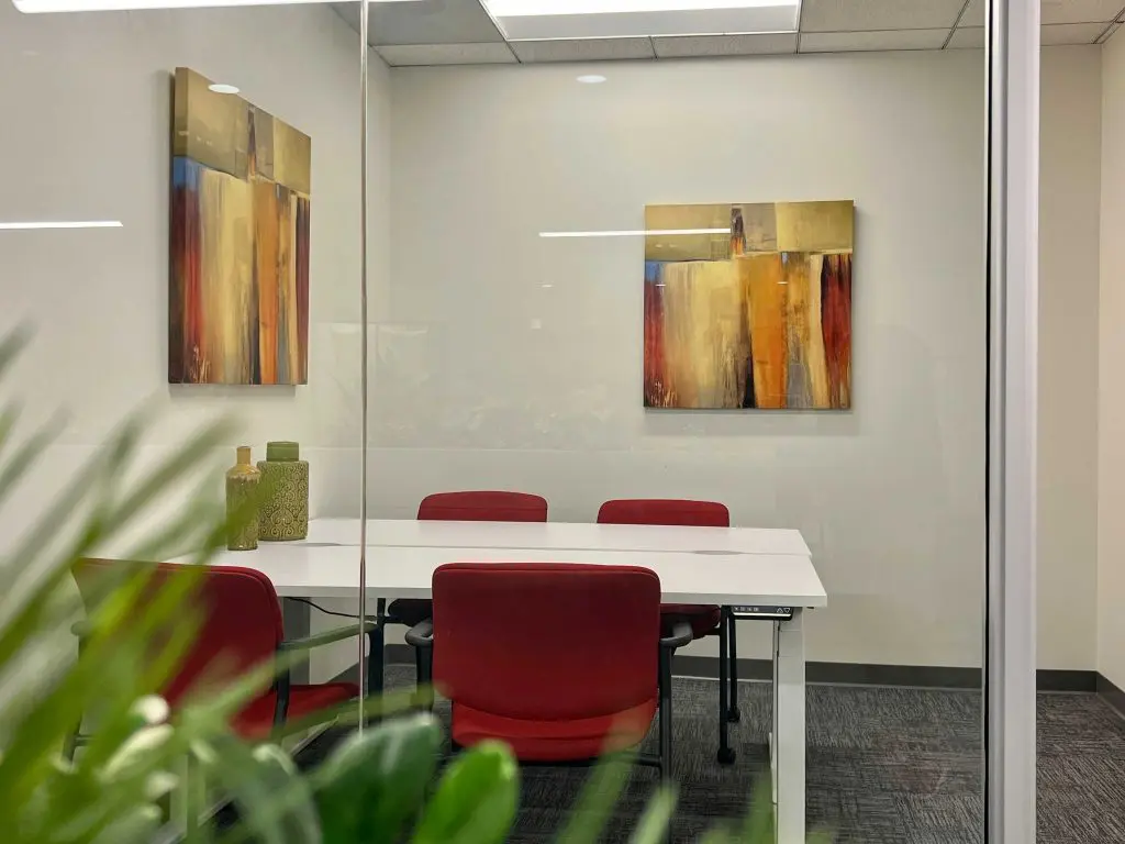 Barrister Suites Office Lease A small conference room with three red chairs around a white table, abstract artwork on the walls, and decorative vases on the table; green plant leaves are visible in the foreground.