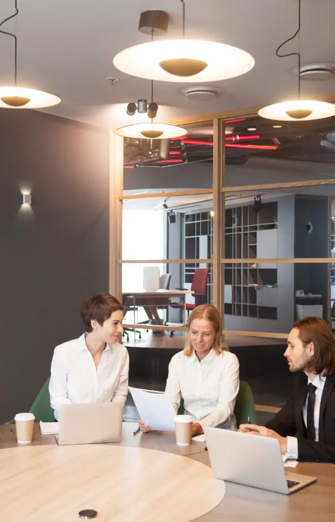 Barrister Suites Office Lease Three people in business attire sit at a round table with laptops and coffee cups, having a discussion in a modern office with glass walls and ceiling lights.