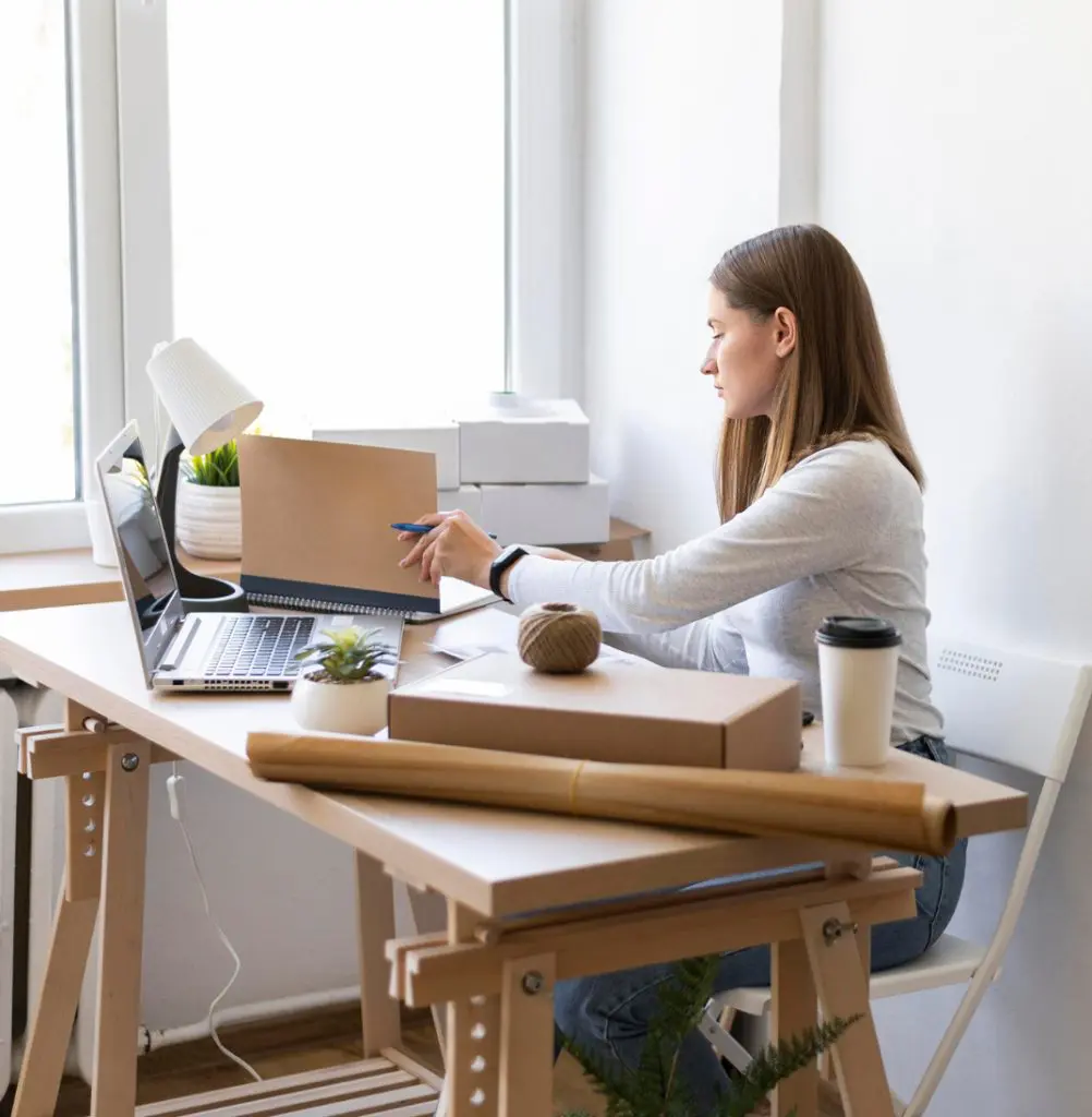 Barrister Suites Office Lease A woman sits at a desk with a laptop, boxes, and packing materials, preparing a package. She writes on a cardboard box, surrounded by a coffee cup, lamp, and small plant near a bright window.