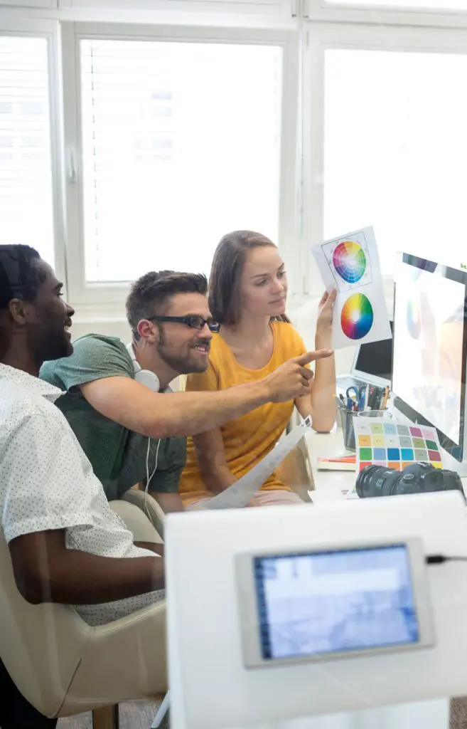 Barrister Suites Office Lease Three people sit at a desk in an office, looking at a computer. One person points at a printout of a color wheel held by another, while the third person watches, all discussing color choices for a project.