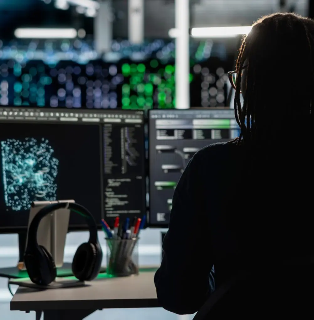 Barrister Suites Office Lease A person with braided hair works at a desk with two monitors displaying data and graphics. Headphones hang on the monitor, and colorful lights blur in the background, suggesting a tech or cybersecurity setting.