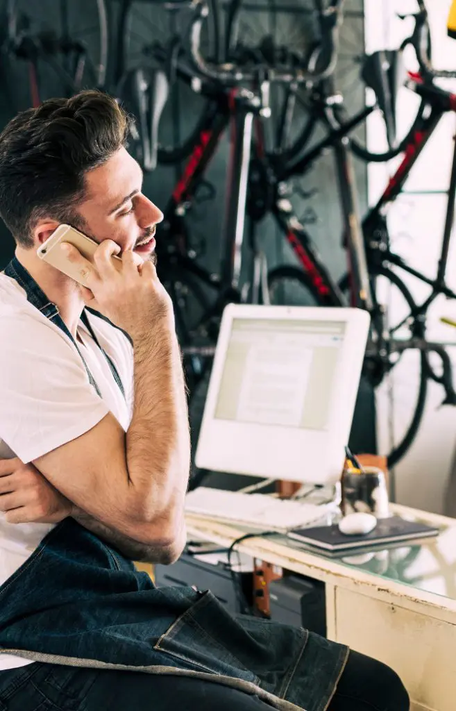 Barrister Suites Office Lease A person wearing an apron smiles while talking on a smartphone in a bike shop, with bicycles hanging on the wall and a computer on the desk nearby.